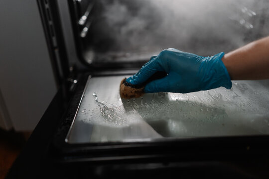 Close-up view of cleaning company worker wearing blue glove using sponge for cleaning glass door of gas oven in dark kitchen room. Concept of hygiene and cleanliness in domestic environment.