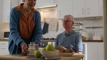 Slow motion of elderly couple preparing for breakfast in the kitchen while careful wife serving table where senior man with smartphone scrolling through online news - Powered by Adobe