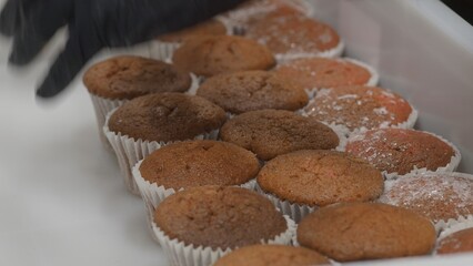 Gloved hand arranging freshly baked chocolate and strawberry muffins in white container, creating a visually appealing display of sweet treats