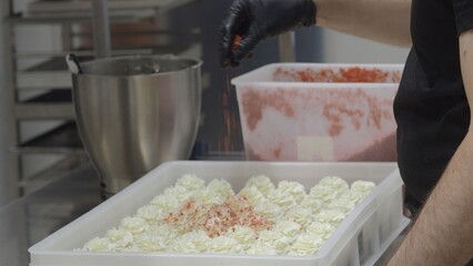 Professional baker wearing black gloves sprinkling red velvet crumbs over white iced cupcakes,...