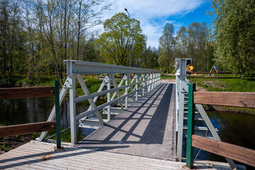 A small pedestrian bridge with crisscrossed metal supports spans a waterway, surrounded by trees and open park space with a playground in the distance.