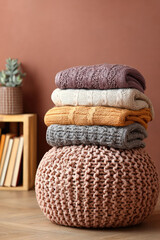 Fototapeta premium A stack of folded knit sweaters rests atop a woven pouf adjacent to a succulent and bookcase