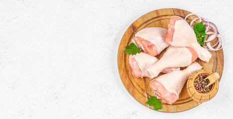 Raw chicken drumsticks arranged on a wooden board with onion rings, parsley, and peppercorns, ready...