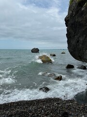 Waves crashing against rocks on a pebble beach with cloudy sky. Suitable for use in travel content,...