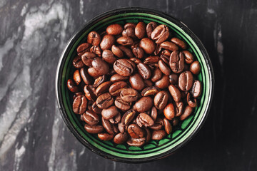 Top view of coffee beans. Brown roasted beans in a green ceramic bowl on black marble background. Morning energy drink ingredient. Natural caffeine stimulant for brewing fresh hot beverage.