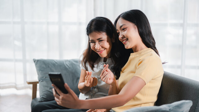 Asian Adult Woman and Older Asian Woman Taking Selfie Together on Mobile Phone on Sofa in Living Room Smiling and Posing Mini Heart Hand Sign At Home