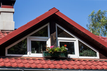 gable attic window with white frame and flower box on red tiled roof of cozy house with blue sky...