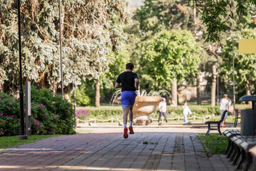 Fototapeta premium man jogging in green city park with fountain and walking people in background, active lifestyle outdoor scene on sunny summer day, urban recreation area with trees, benches and natural surroundings