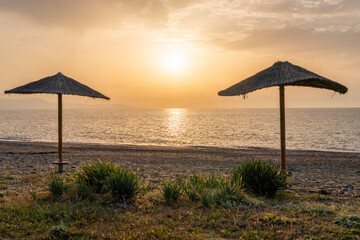 Two straw beach umbrellas against sea at bright sunset. Golden light fills scene, creating a...