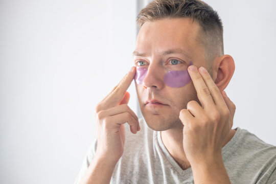 Man applying purple gel under eye patches in front of mirror for skincare routine and freshness. Beauty and skin care concept. Male grooming, facial care routines, anti-aging through under-eye care - Powered by Adobe