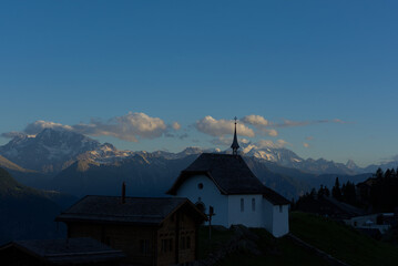 A serene church with a cross-topped steeple stands silhouetted against snow-capped mountains and a twilight sky, creating a peaceful and inspiring alpine scene.