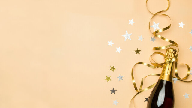 Celebration Flat Lay with Champagne and Stars. A festive flat lay featuring a champagne bottle, a silver streamer, and scattered gold star confetti on a pale peach background, leaving ample copy space