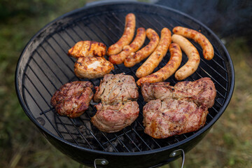 Mixed meats cooking on outdoor charcoal grill. Assorted grilled meats including sausages, chicken, and pork steaks sizzling on a round charcoal barbecue in a backyard setting.