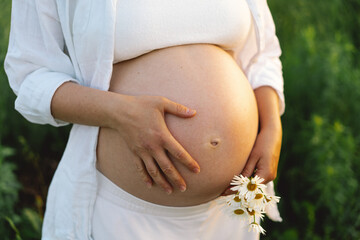 A pregnant woman stands in a green field. She gazes lovingly at her baby bump as the sun sets behind her, creating a serene atmosphere.