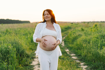 A pregnant woman stands in a green field. She gazes lovingly at her baby bump as the sun sets behind her, creating a serene atmosphere.