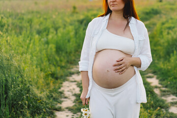 A pregnant woman stands in a green field. She gazes lovingly at her baby bump as the sun sets behind her, creating a serene atmosphere.