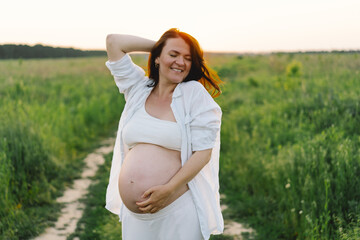 A pregnant woman stands in a green field. She gazes lovingly at her baby bump as the sun sets behind her, creating a serene atmosphere.