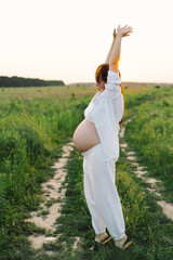 A pregnant woman stands in a green field. She gazes lovingly at her baby bump as the sun sets behind her, creating a serene atmosphere.