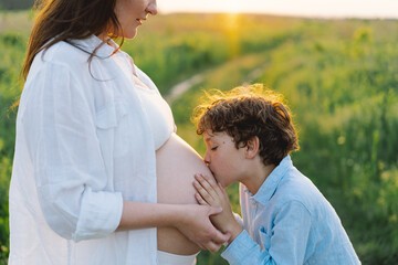 A pregnant woman stands in a lush green field, gently holding her baby bump. Her son leans against her, both sharing a tender moment amidst tall grass and a warm sunset.
