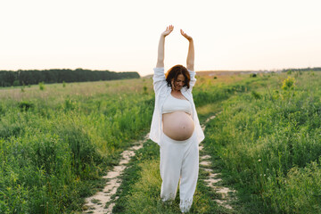 A pregnant woman stands in a green field. She gazes lovingly at her baby bump as the sun sets behind her, creating a serene atmosphere.