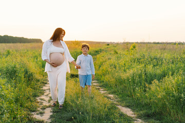 A pregnant woman stands in a lush green field, gently holding her baby bump. Her son leans against her, both sharing a tender moment amidst tall grass and a warm sunset.