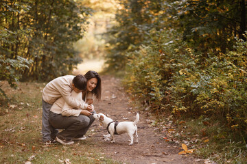 Side view of an Asian mother and her young son laughing while walking a small white dog on a forest trail. They wear casual autumn clothes. Great for family, parenting, and outdoor lifestyle themes.
