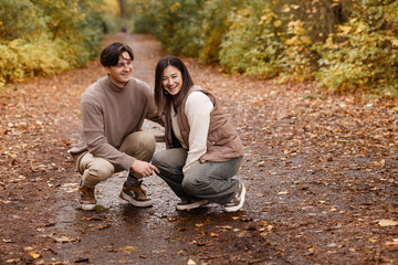 Side view of an Asian couple laughing while walking a small white dog on a forest path. Casual autumn clothes. Great for love, outdoor lifestyle, pet owners, and relationship themes. Copy space.