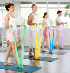 Slender young men and women doing arm and shoulder workout with resistance band in standing...