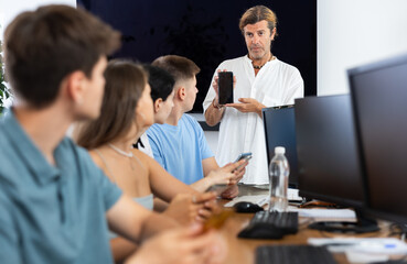 At computer education class, male teacher stands with mobile phone in hands and explains about...