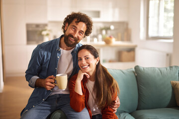 A couple sits comfortably on a couch in their living room, smiling and holding a warm drink. The afternoon sunlight brightens the space, creating a welcoming atmosphere.