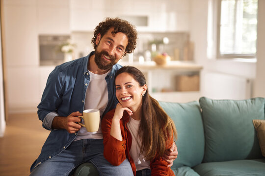 A couple sits comfortably on a couch in their living room, smiling and holding a warm drink. The afternoon sunlight brightens the space, creating a welcoming atmosphere.
