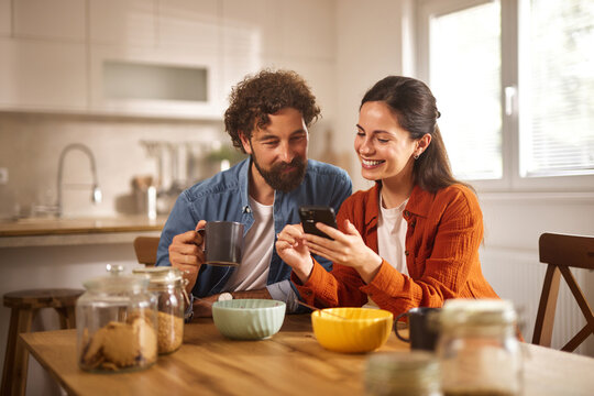 A couple sits at a wooden table in their home office, smiling and sharing a moment while looking at a smartphone. They have coffee mugs and bowls of snacks in front of them.