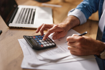 A person uses a calculator and notepad while working at a desk with a laptop, focusing on detailed...