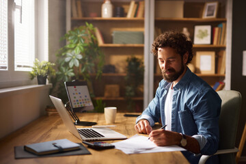 A man sits at a wooden desk in a well-lit home office, diligently writing notes while using a...