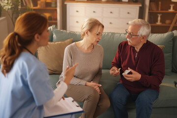 In a living room, a female home nurse in scrubs reviews a medication plan with an older couple. The man holds a pill blister pack as the nurse discusses treatment details with the couple.