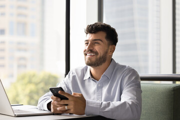 Great news. Overjoyed excited young businessman working online sit by notebook hold phone look away...