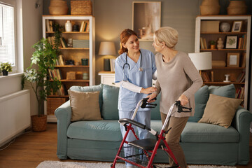 In the living room, a friendly home healthcare nurse in scrubs assists an elderly woman who uses a...