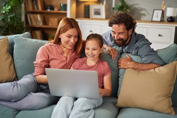 Parents and their daughter share joyful moments while watching something together on a laptop,...