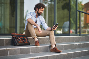 A man in business attire enjoys a moment of relaxation on outdoor steps, using his smartphone while...