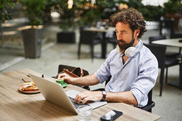 A man is focused on his laptop while enjoying coffee and a pastry at a contemporary cafe. The environment features greenery, creating a productive workspace.
