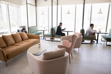 Comfy collaborative workspace. Open coworking space interior. Relaxing lounge zone with soft couch armchairs low table. Diverse people working on notebooks at individual desks with seats in background