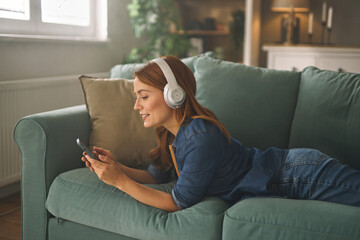 A woman with long red hair lies comfortably on a couch, wearing headphones and smiling as she uses a smartphone in a warmly lit living room.