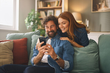 A man and woman sit closely on a comfortable couch, looking at a smartphone together, sharing smiles in a cozy living room filled with books and plants.