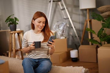A woman sits comfortably on a couch, sipping coffee from a mug, and looking at her phone. Around...