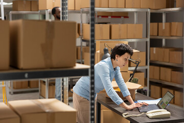 Young woman at her desk reviewing shipment data for delivery services, verifying taped boxes in a...
