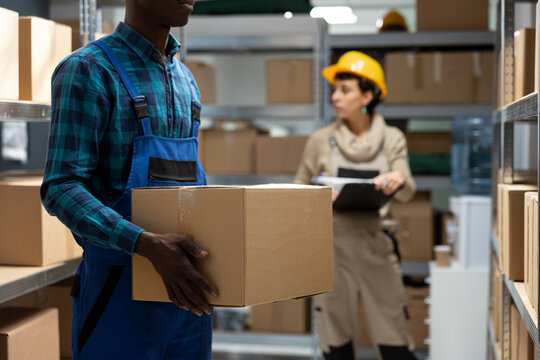 Industrial depot environment focused on import export efficiency and storage. Black employee handles manual work tasks such as inventory tracking, goods handling and shipment preparation.