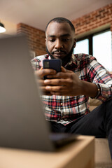 Black male remote worker taking break from laptop, using cellphone to browse social media and stay updated. Young african american professional balancing digital tasks and relaxation in home office.