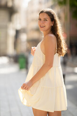 Cheerful girl in white dress walking along the street among architecture with a bag