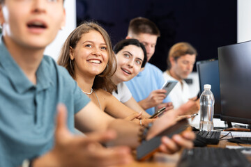 Cheerful young female student studying together with coursemates in computer workshop, actively using mobile phone to access online learning materials or participating in group discussions
