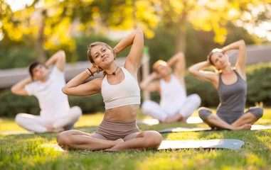Selbstklebende Fototapeten Fit young girl enjoying group yoga session in peaceful environment of verdant summer park, performing stretches in lotus position while sitting on exercise mat © JackF